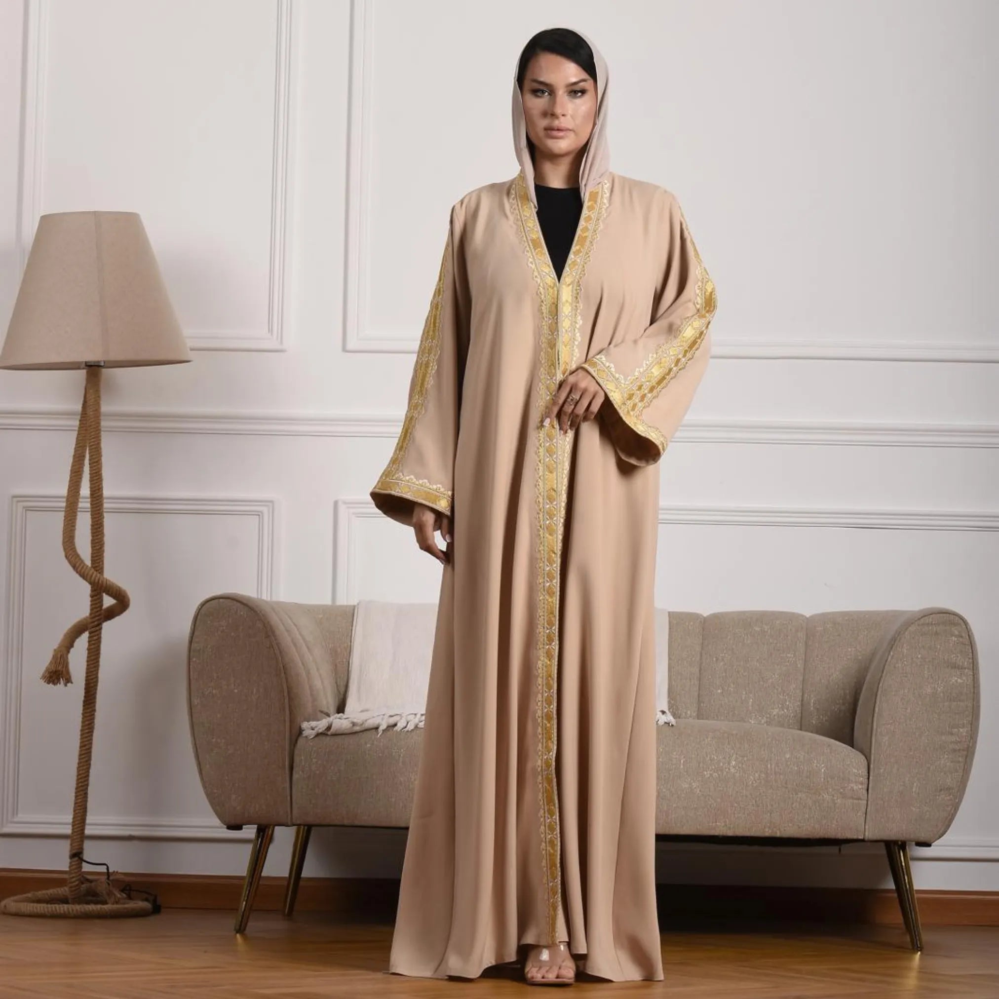 Woman in a beige abaya with gold embroidery standing in a room with a lamp and sofa.