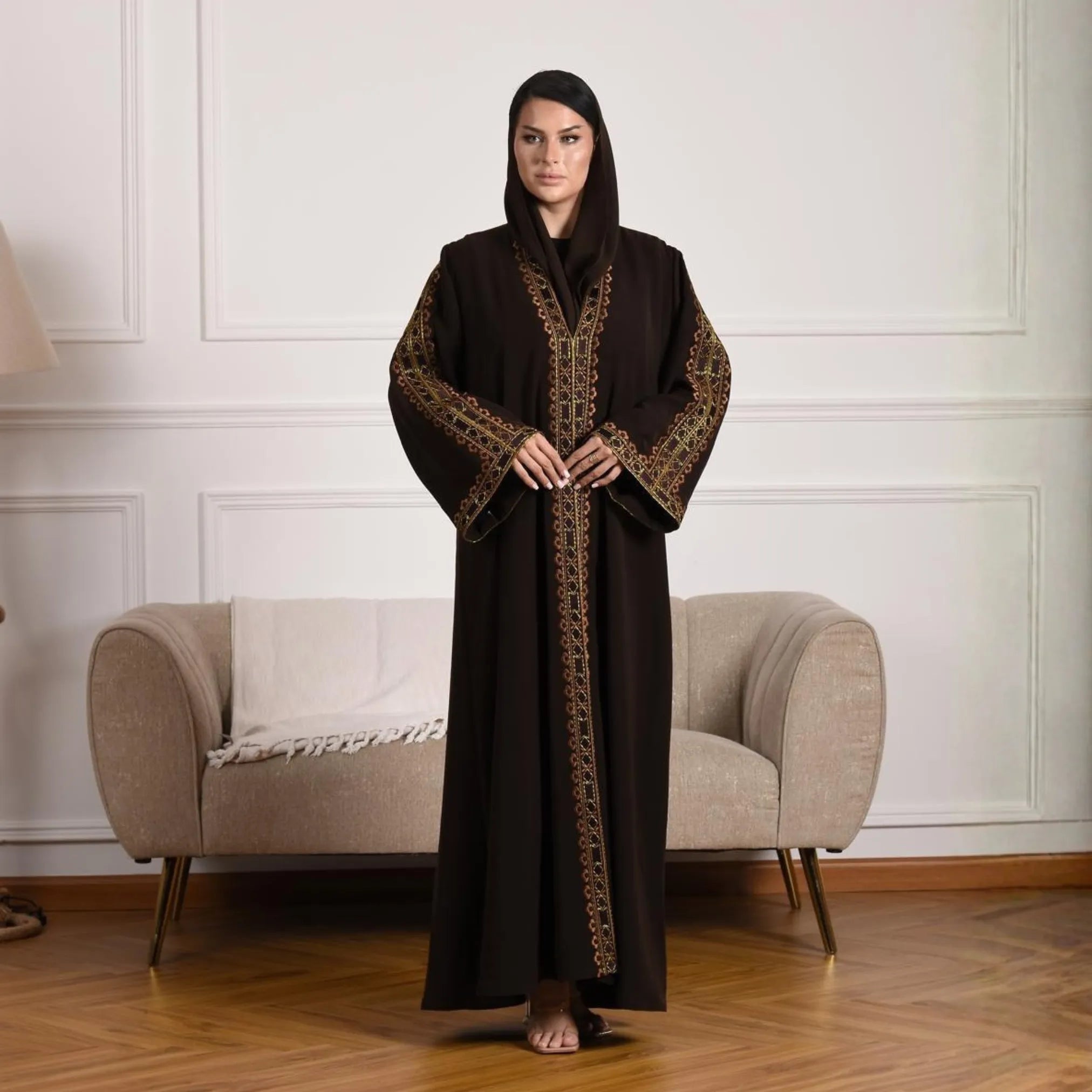 Woman wearing a dark brown abaya with intricate patterns in a room with a beige armchair.