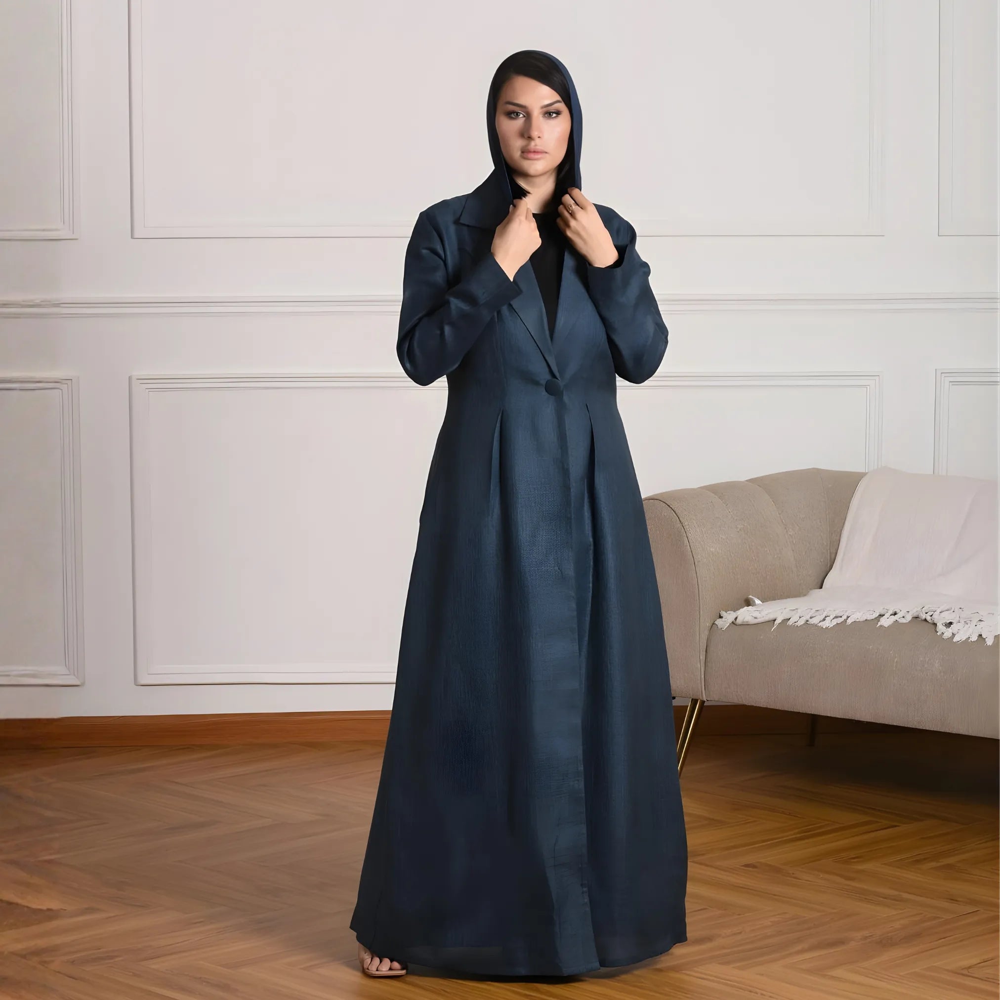 Woman wearing a long dark blue abaya standing in a room with a beige sofa and white walls.