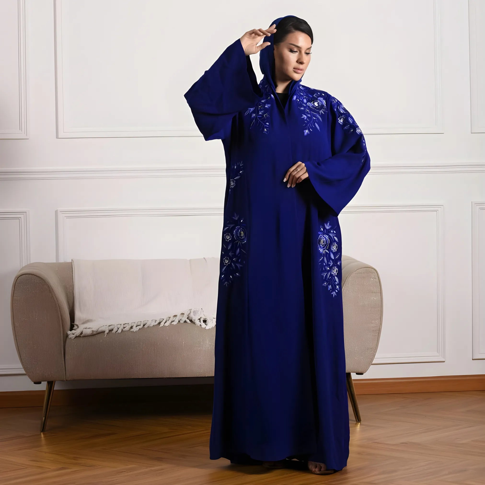 Woman in a blue embroidered abaya standing in a room with a beige armchair.