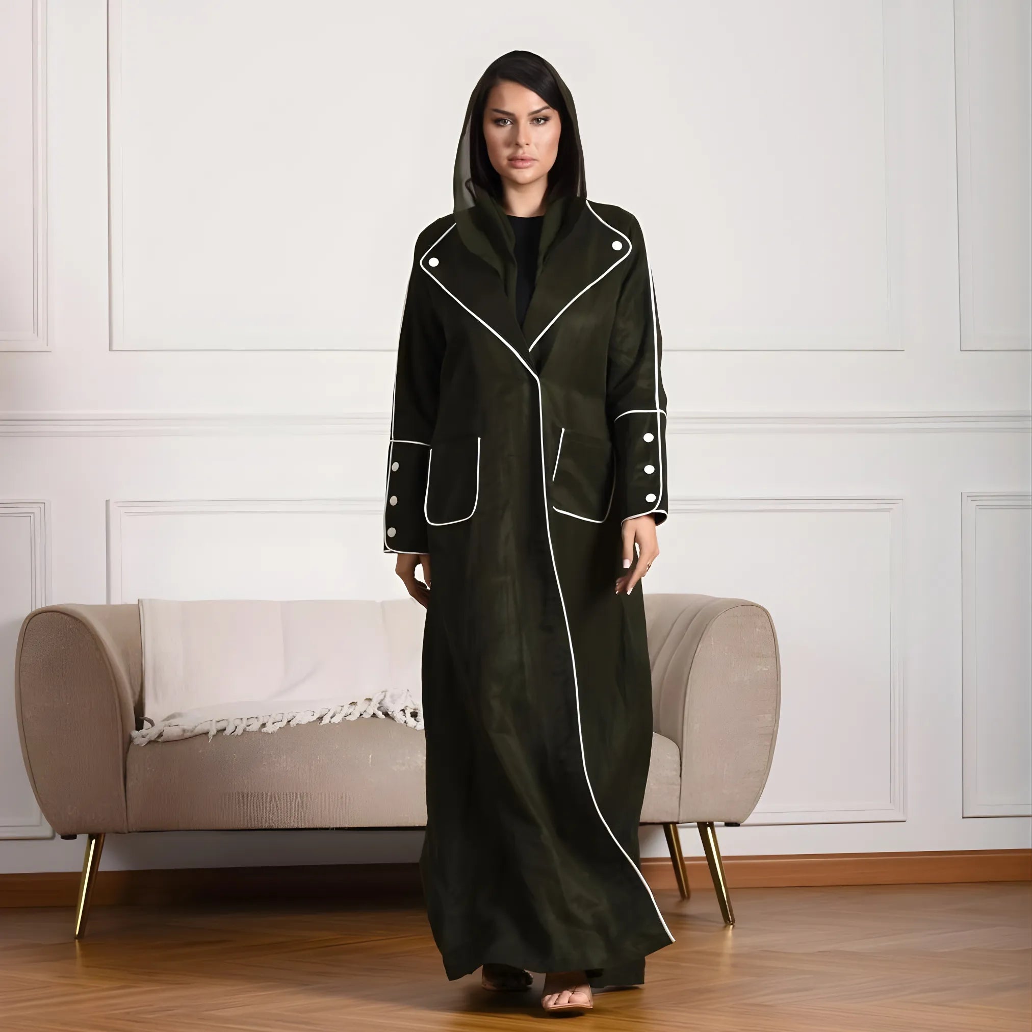 Woman wearing a long dark green coat Abaya with white trim in a room with a beige sofa.