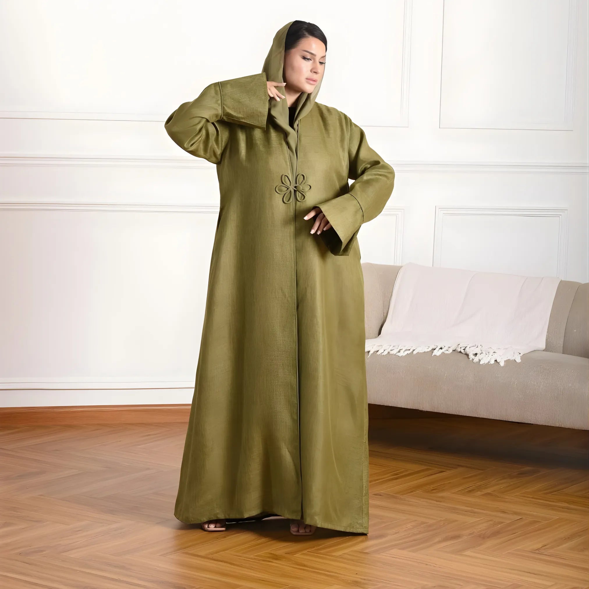 Woman wearing a long olive green coat in a room with a white wall and beige sofa.