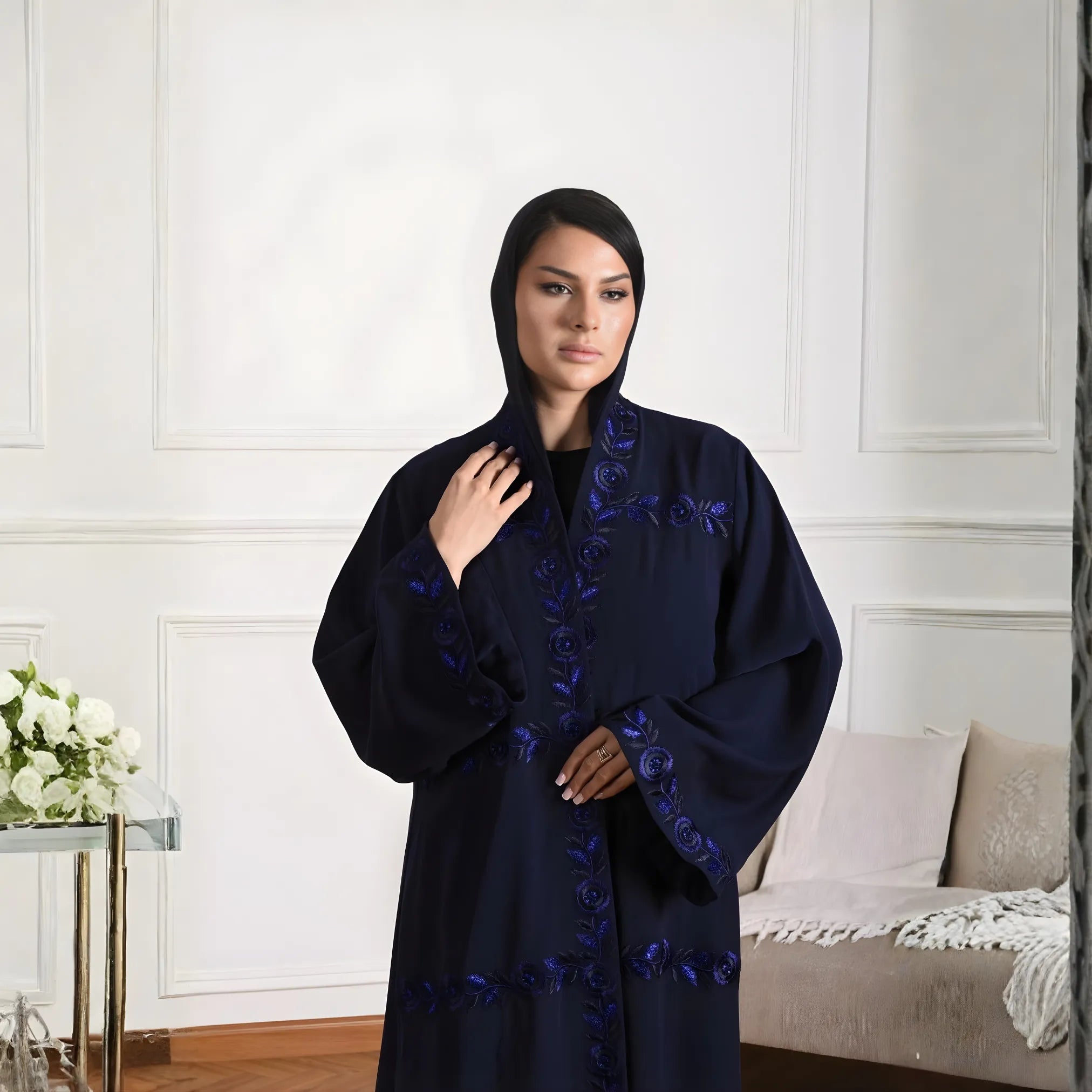 Woman wearing a dark blue abaya with floral embroidery in a room with white walls and a beige sofa.