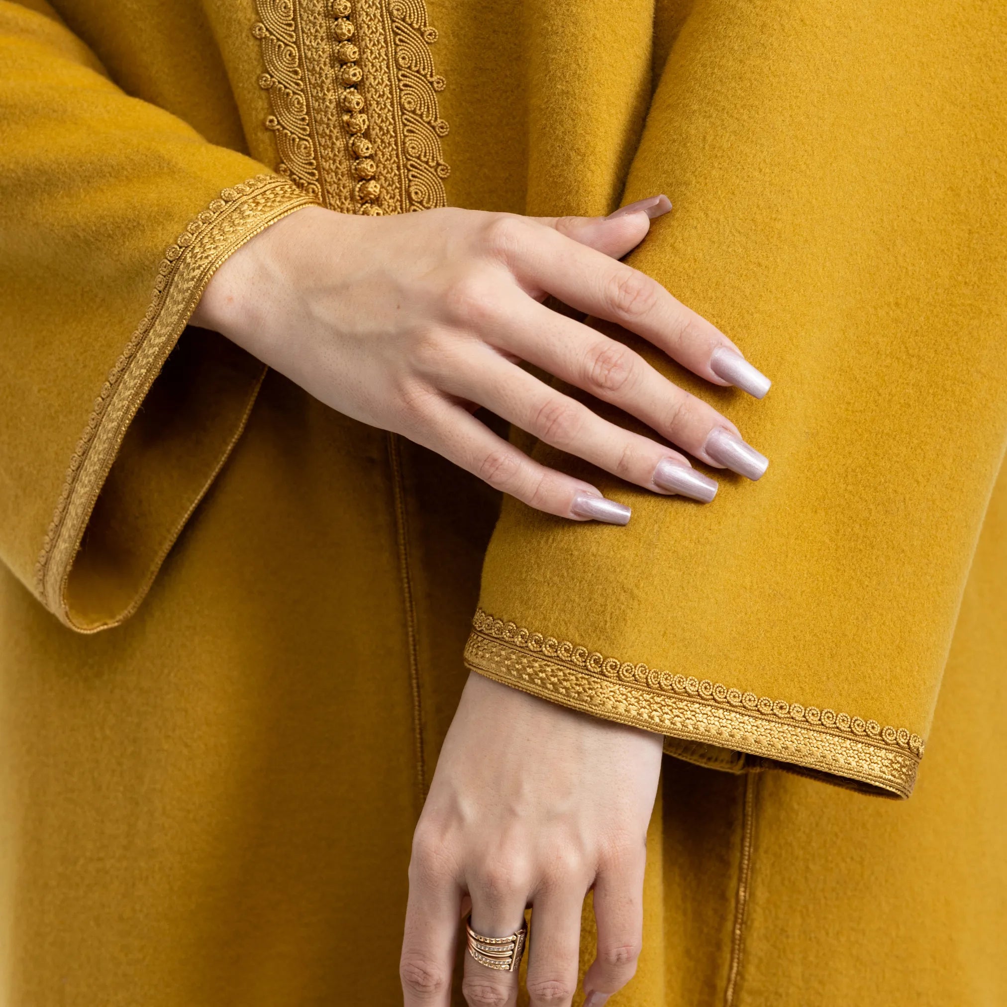 Close-up of a mustard yellow jalabiya with gold embroidery and a hand touching the fabric.