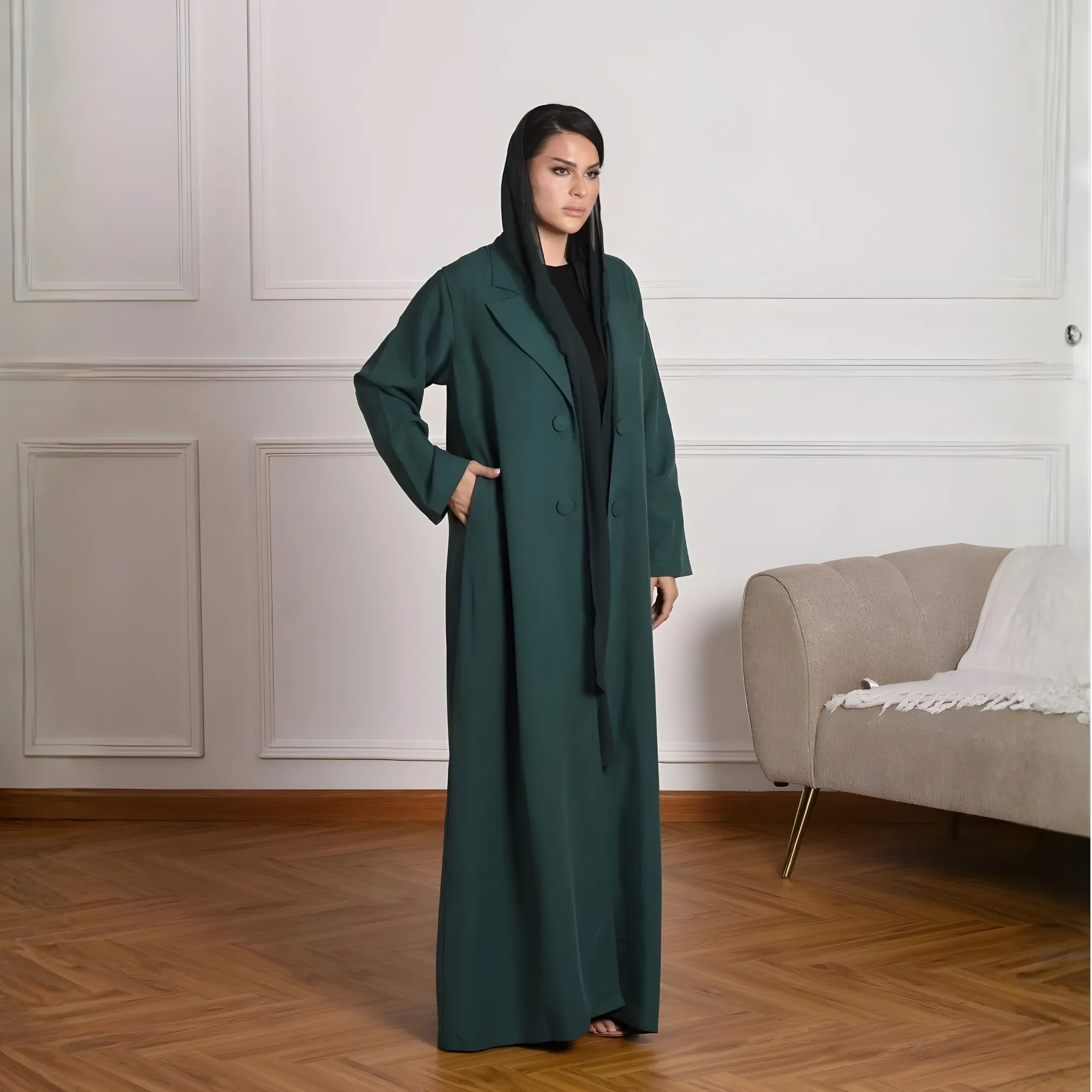Woman wearing a long dark green abaya in a room with a beige sofa and white walls.
