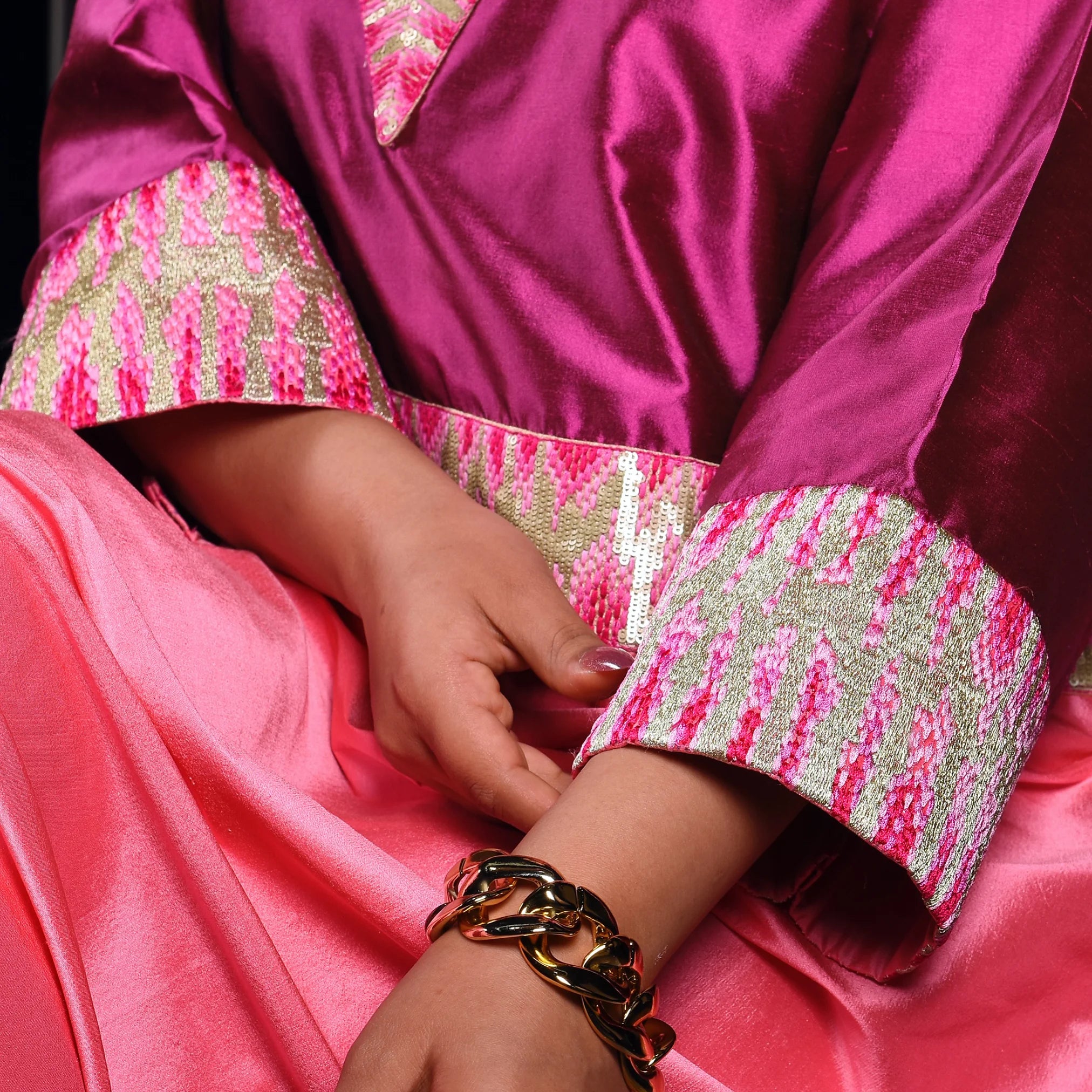 Close-up of a person wearing a pink jalabiya with gold and pink patterned sleeves and a gold bracelet.