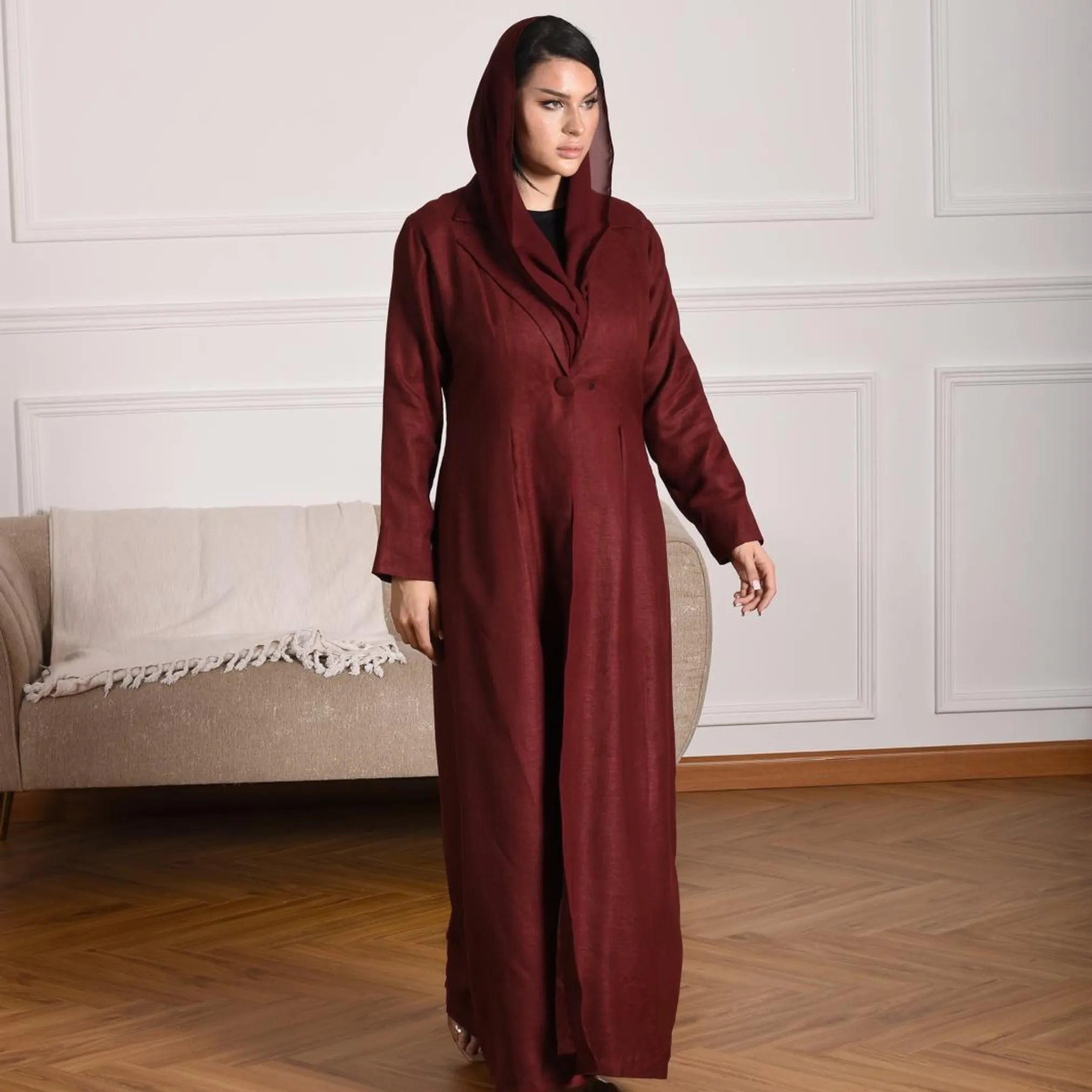 Woman wearing a long burgundy abaya standing in a room with a beige sofa and white walls.