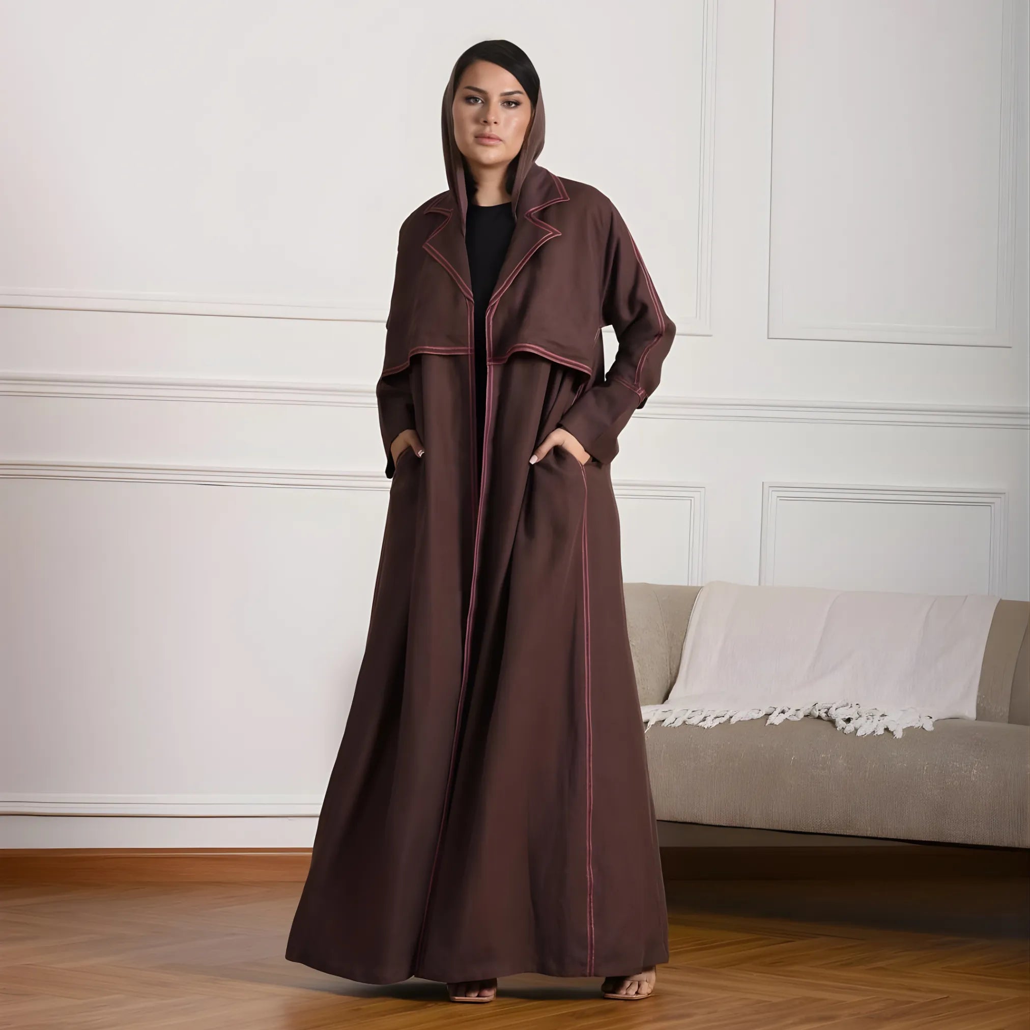 Woman wearing a long brown coat Abaya in a room with a white wall and beige sofa.
