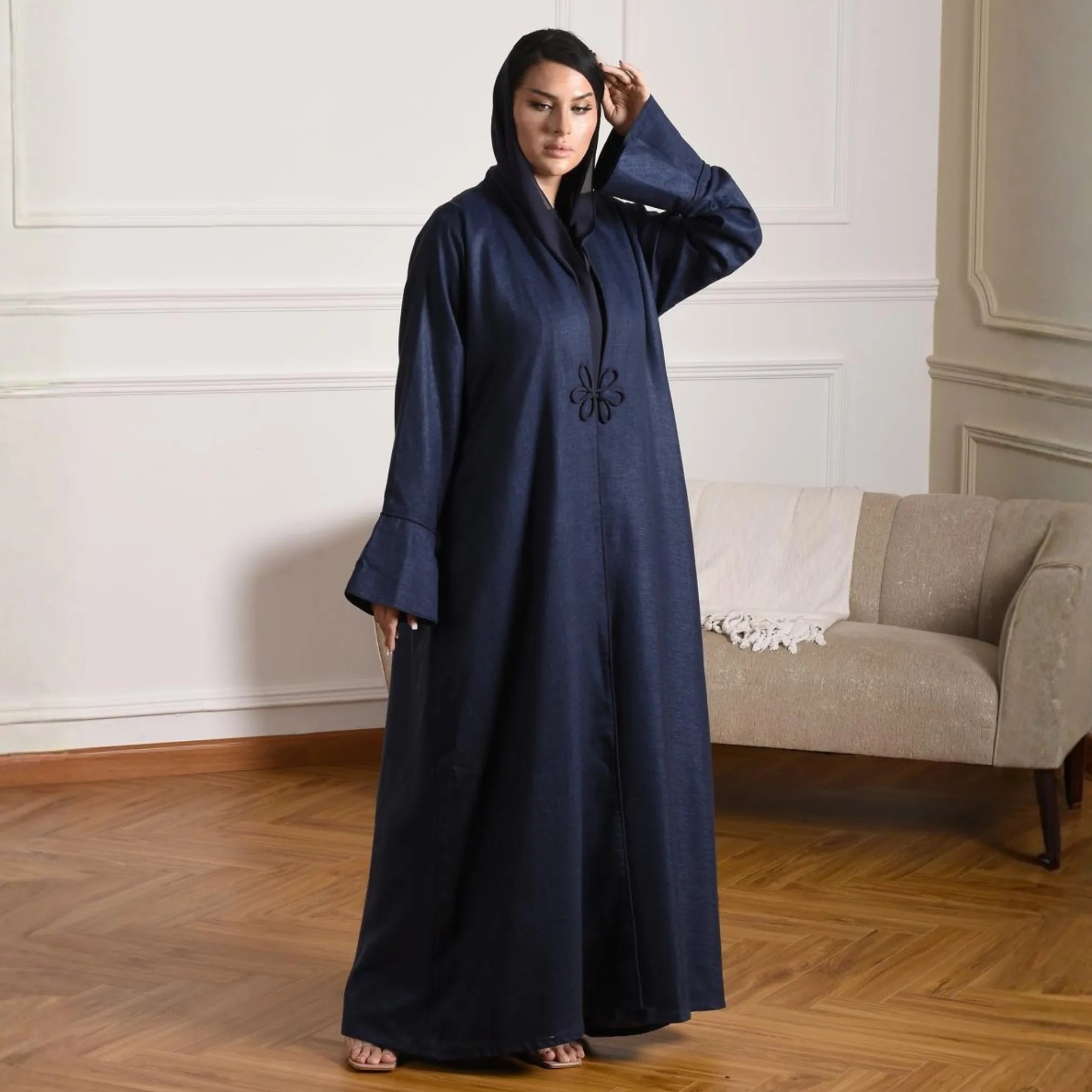 Woman wearing a navy blue abaya with embroidery in a room with a beige sofa and wooden floor.